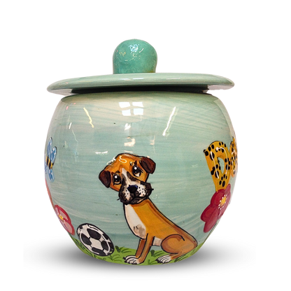 A round, hand-painted treat jar with a pastel blue lid, featuring a charming pup and colorful paw prints amidst a backdrop of grass and soccer balls