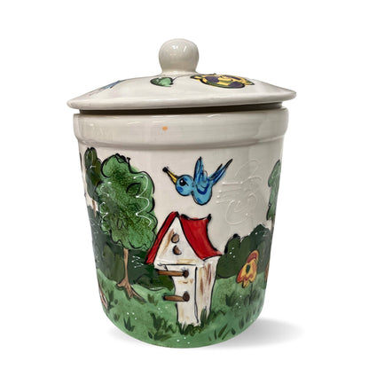 Artisanal ceramic jar with a painting of a blue bird flying towards a red and white birdhouse, surrounded by green bushes and a floral accent