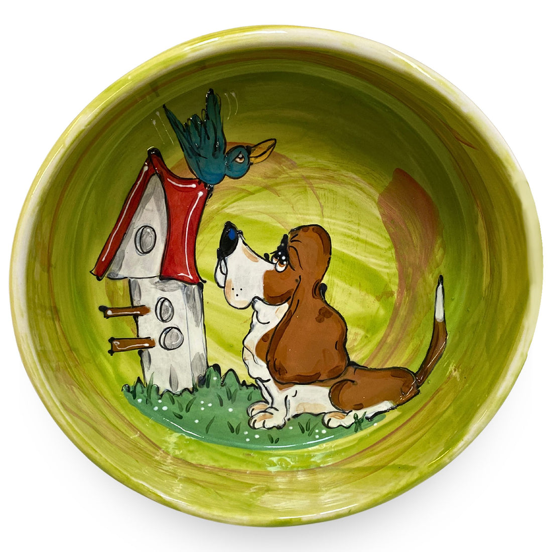 Hand-painted ceramic bowl with a playful basset hound looking at a bird by a red and white birdhouse, against a vibrant green background