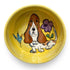 A bright yellow hand-painted ceramic bowl with a basset hound, a blue bird, and a purple flower, evoking a cheerful, sunlit garden atmosphere.