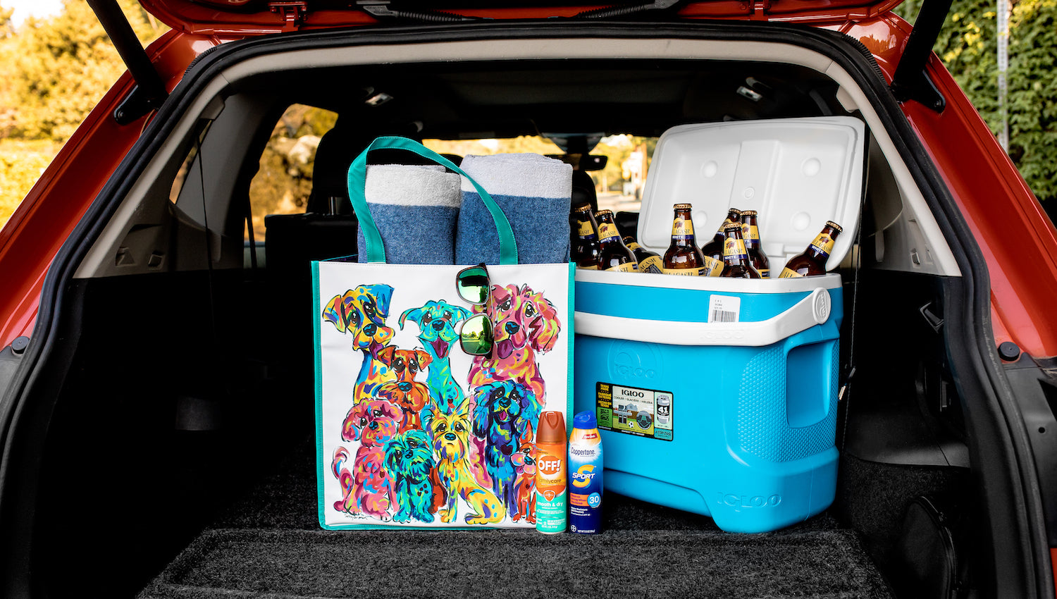 Open car trunk packed for an outing with a colorful vinyl tote featuring vibrant dog artwork by Debby Carman, blue and white towels, a blue Igloo cooler filled with bottled beverages, and sun protection products.