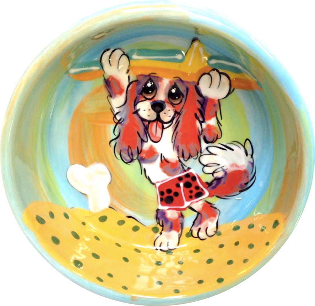 image of cavalier King Charles spaniel taking surfboard to the waves in a sunset beach scene custom created pet bowl hand painted by faux paw productions artist Debby Carman 